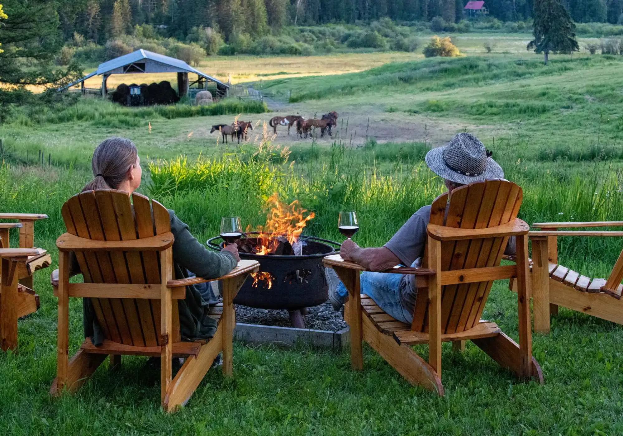 Two people in wooden chairs beside a fire bowl at Kayanara, looking out over the meadow toward grazing horses.