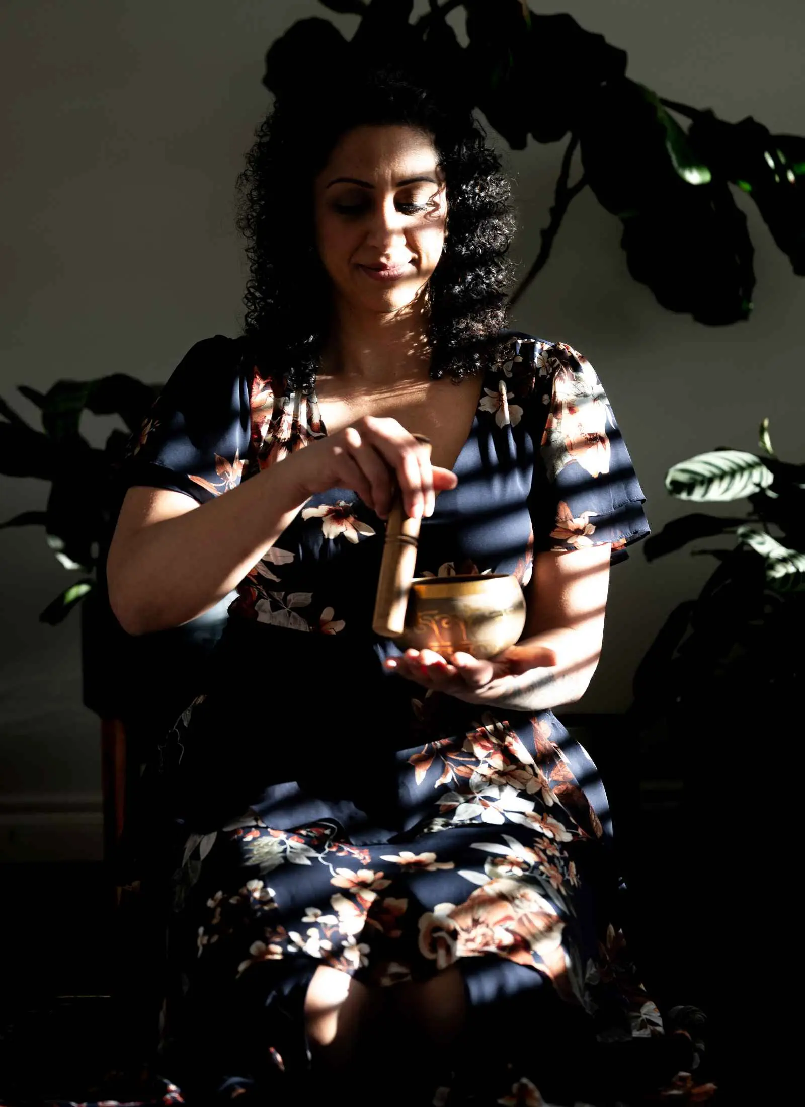Pam Dhillon seated in dappled light, holding a singing bowl.