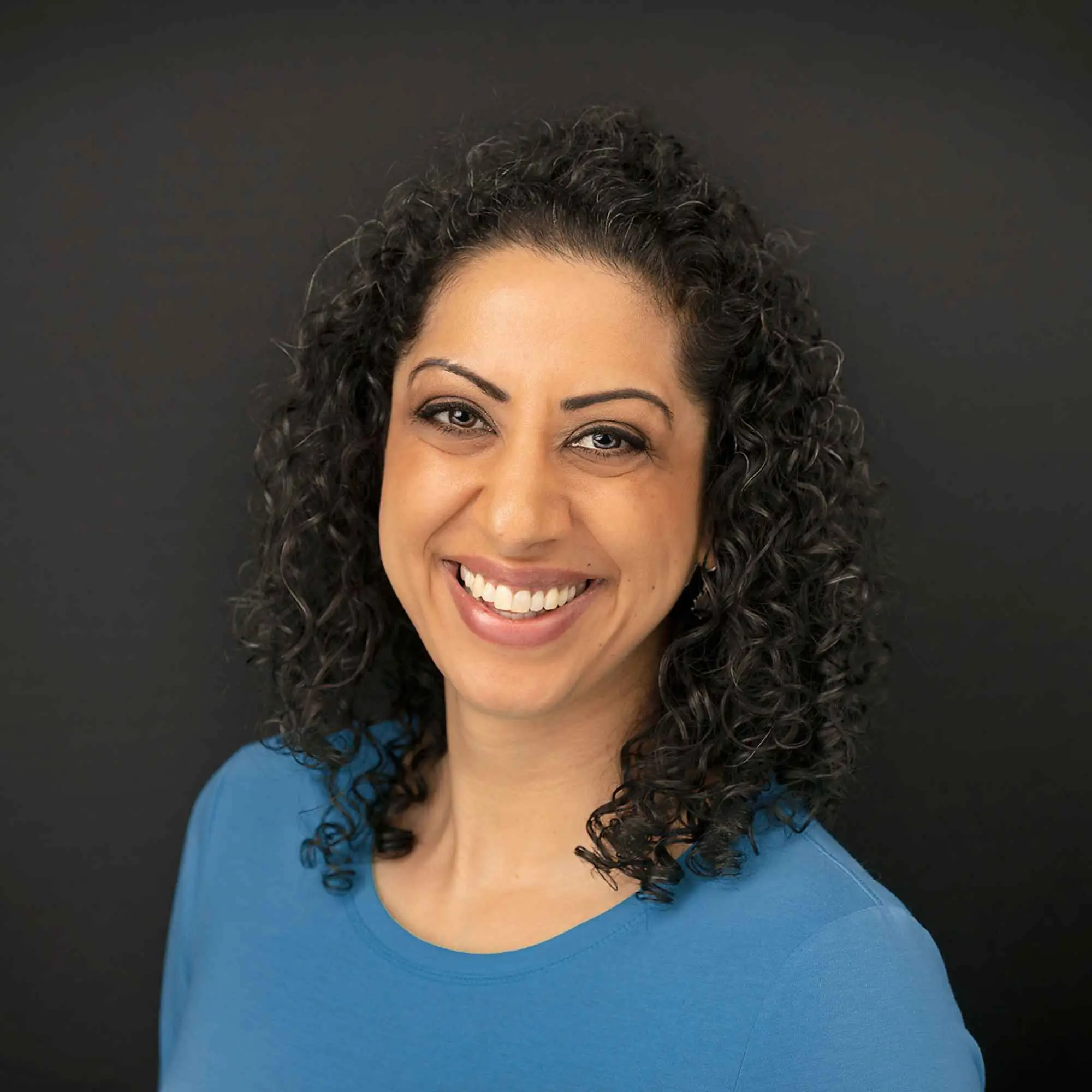 Pam Dhillon, smiling, against a dark studio background.