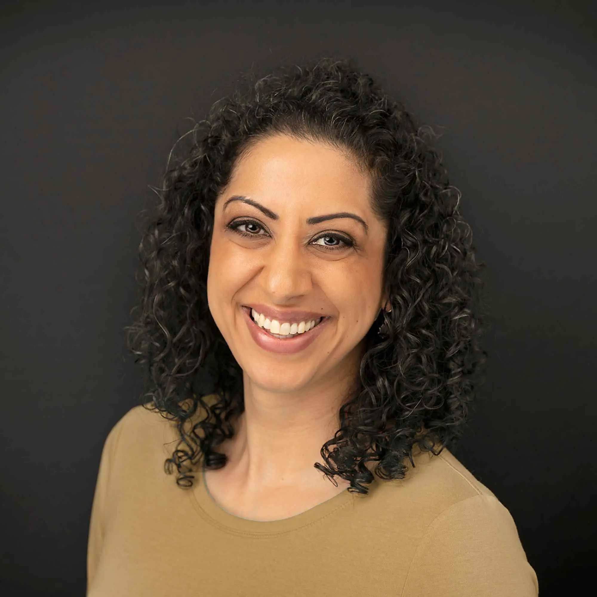 Pam Dhillon, smiling, against a dark studio background.