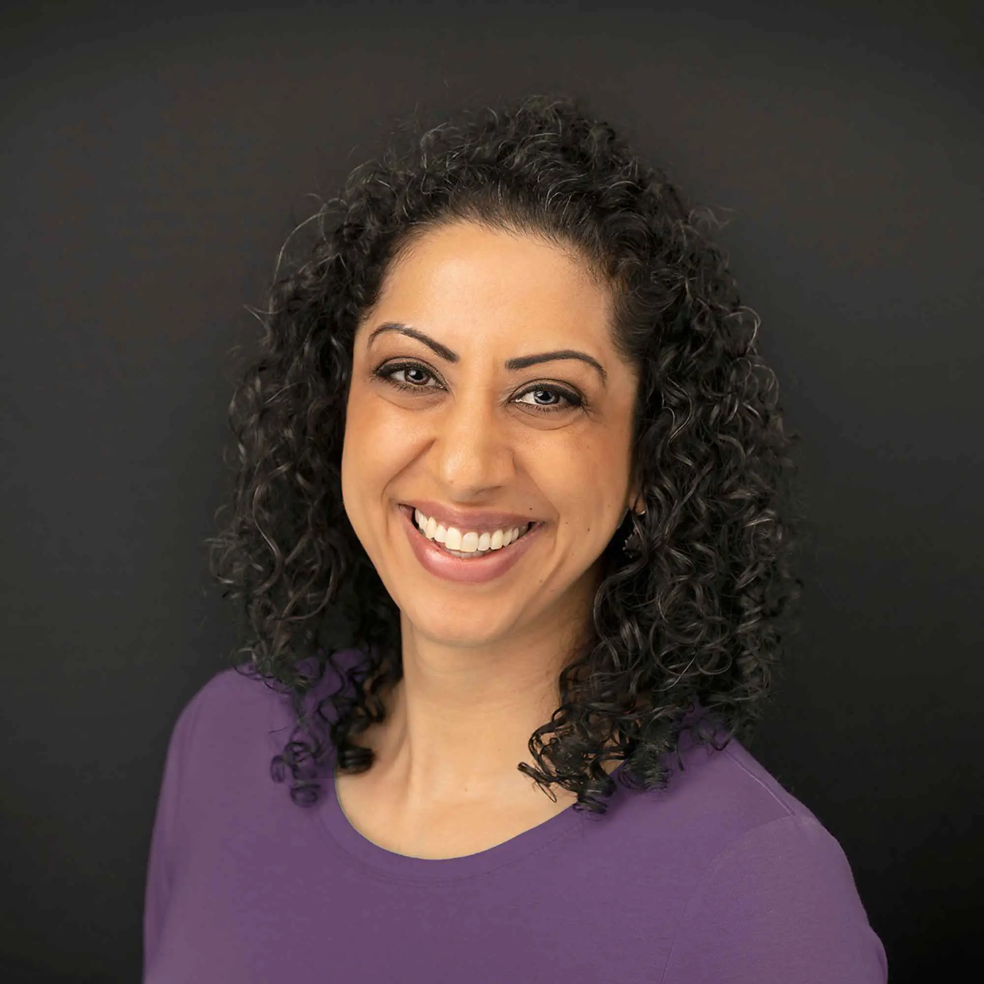 Pam Dhillon, smiling, against a dark studio background.