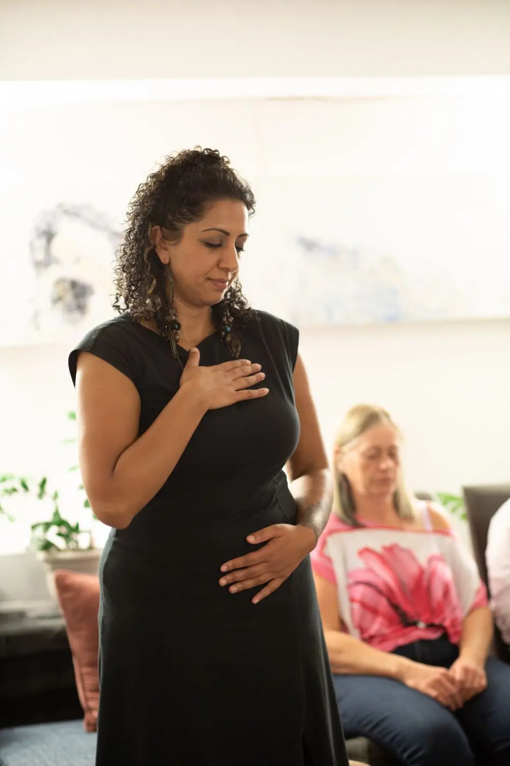 Pam Dhillon standing with one hand on her heart and one on her belly, eyes closed, in a bright room with others seated nearby.