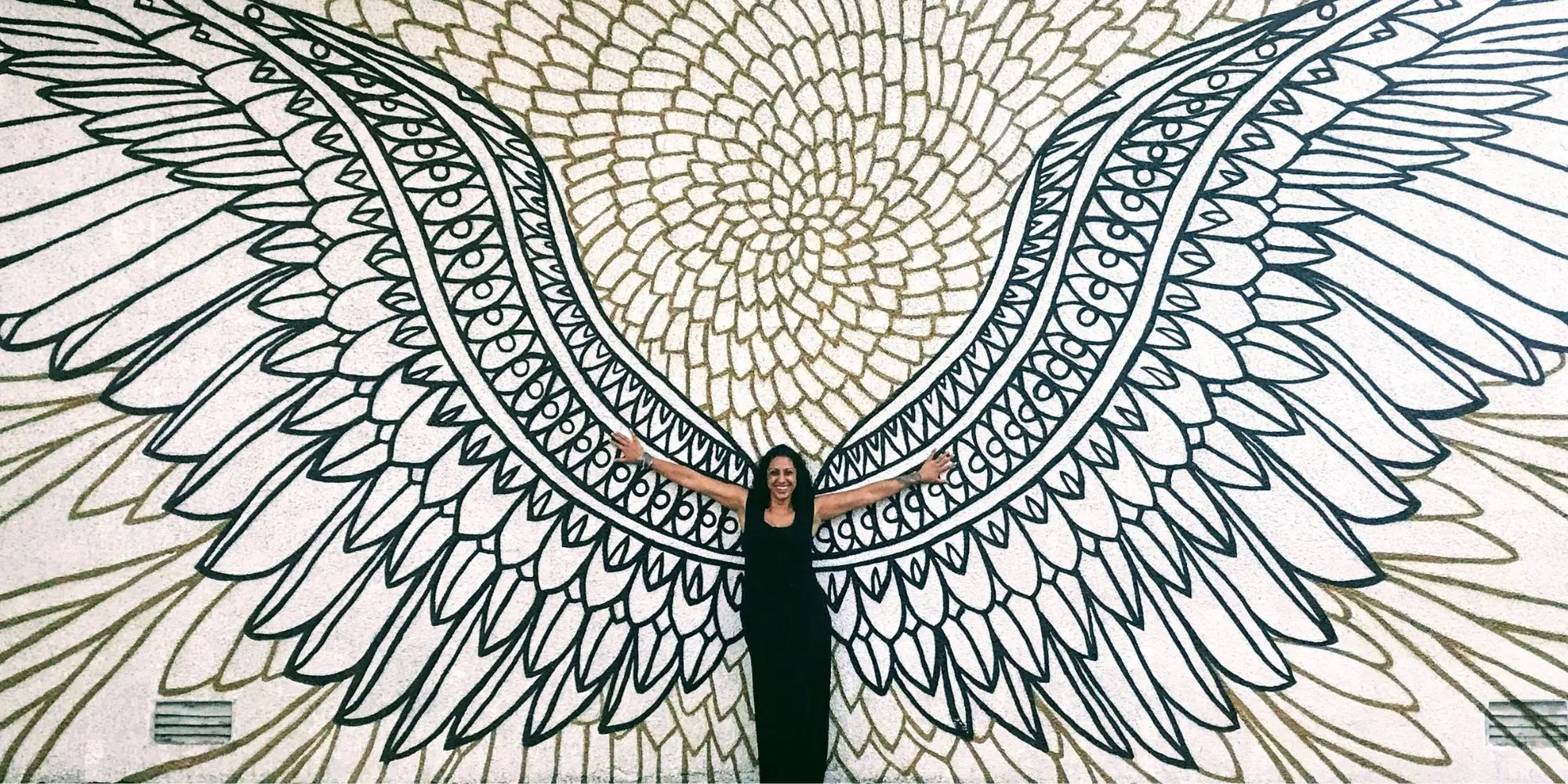 Pam Dhillon standing with arms outstretched in front of a large painted mural of white wings.