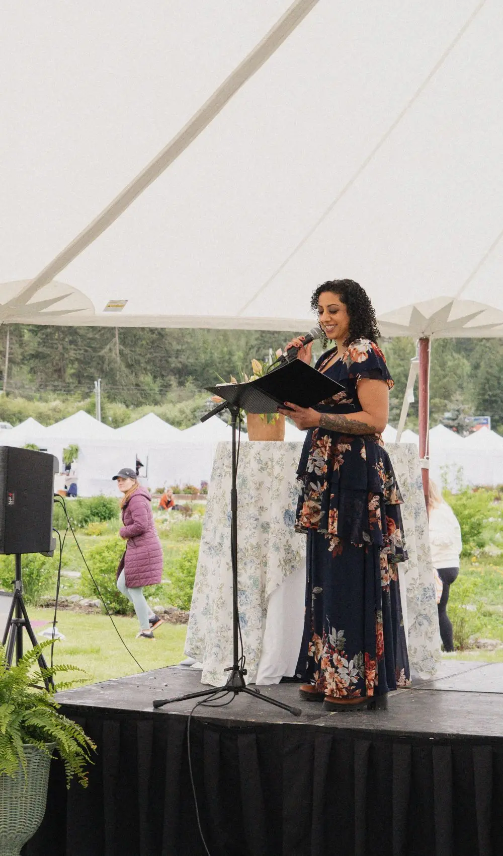 Pam Dhillon at a lectern on an outdoor stage under a white tent, with market stalls and trees behind.