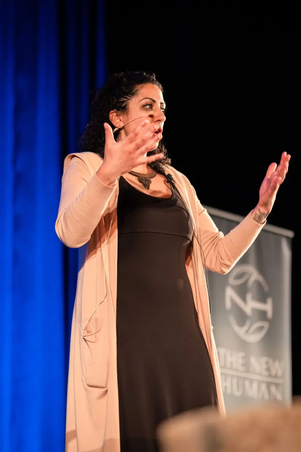 Pam Dhillon speaking on stage at a New Human event, hands raised mid-sentence, a blue curtain behind her.