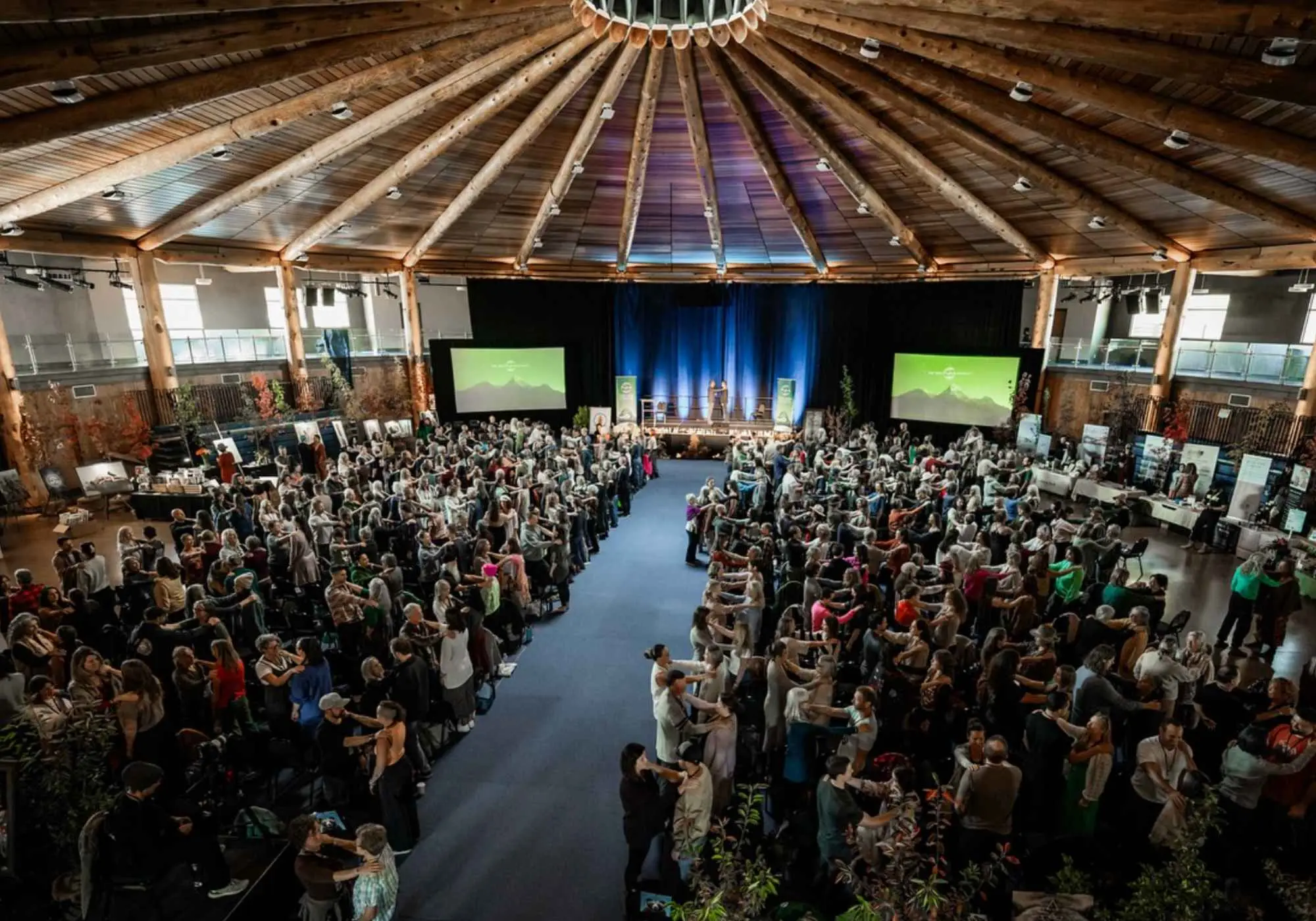 A packed timber hall at a New Human event, hundreds of people standing in pairs while two figures speak from the lit stage.